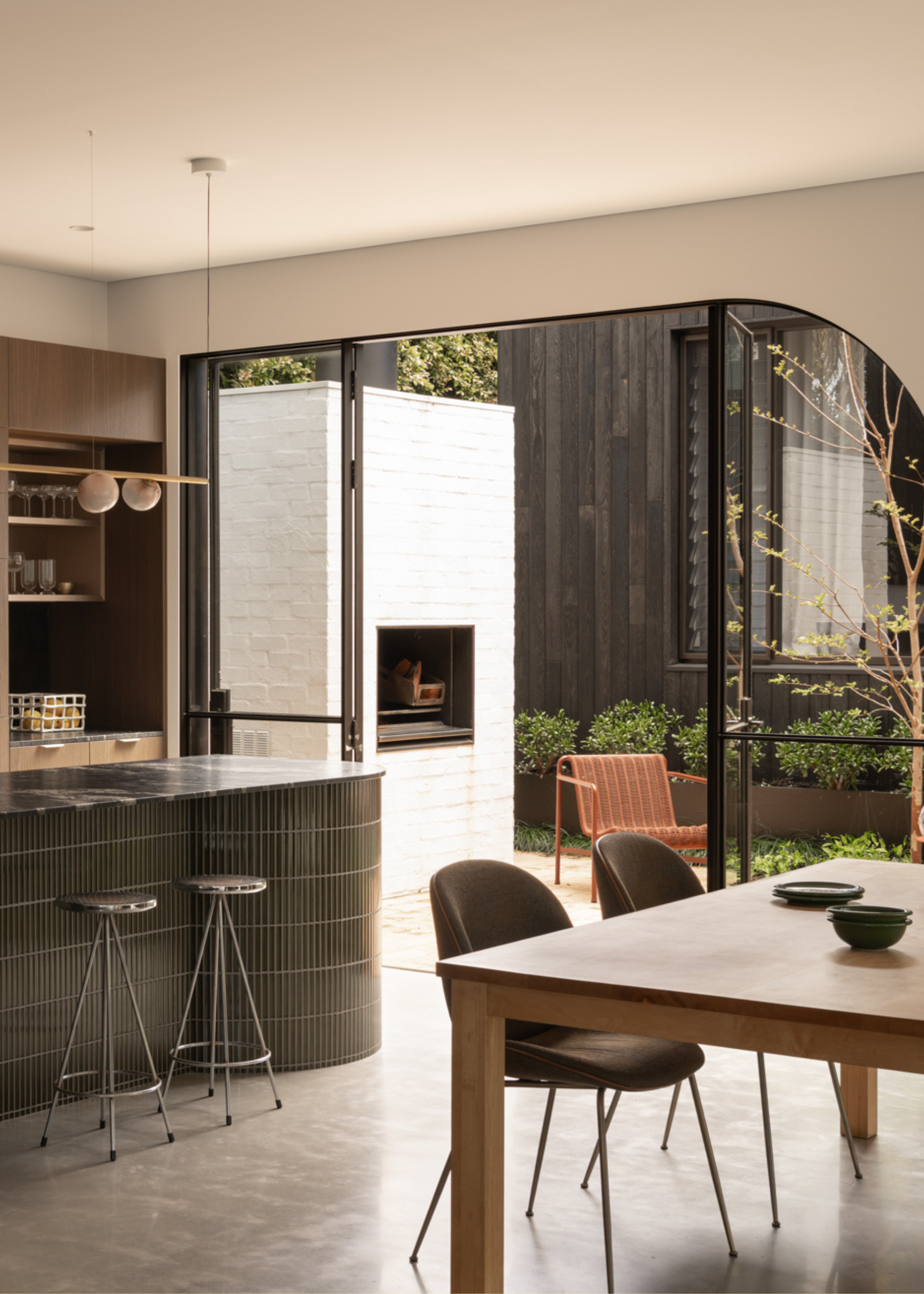 A bright open concept kitchen and dinding room with a wood table, ceramic bowls, brown chairs, a tiled island, chrome stools, and a door to a backyard