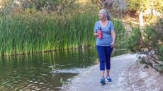 Woman walks beside body of water, holding a water bottle