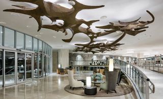 Interior view of Gran Melia, Rio de Janeiro, Brazil featuring light coloured flooring, white, grey and green chairs, round tables, lamps, glass doors and windows, a ramp and a white ceiling with translucent, curved objects and an irregular shaped brown structure suspended from it