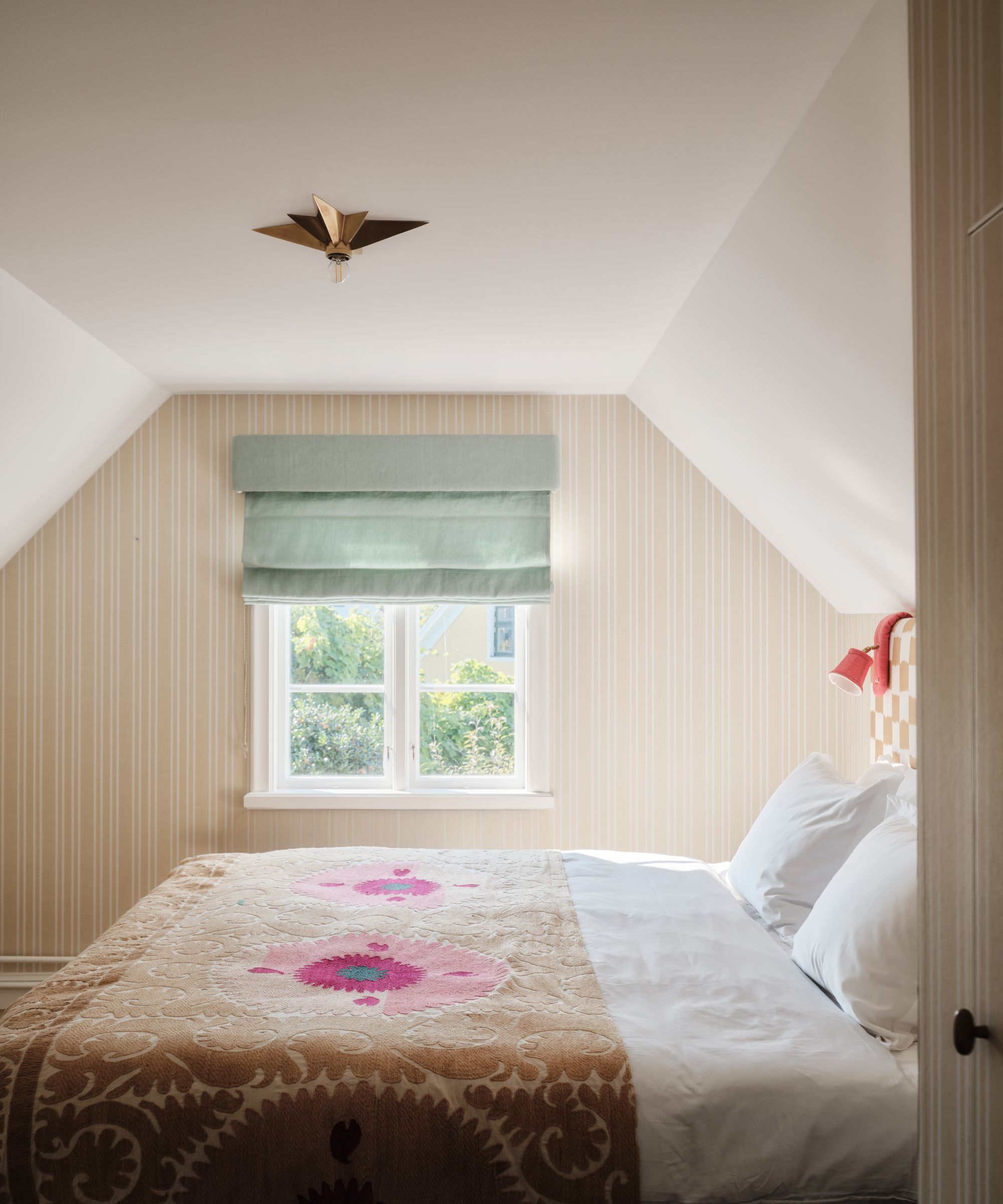 An attic bedroom with a white ceiling and pale yellow paneled walls. A small window has a pale blue Roman blind and the bed is made with white sheets and a pink and yellow patterned throw blanket