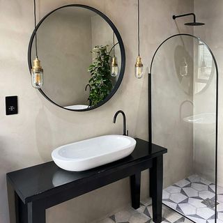 Bathroom with cream coloured concrete walls, rounded glass shower panel with black rim, matching circular mirror, a black vanity with long and shallow sink and two gold and black pendant lights