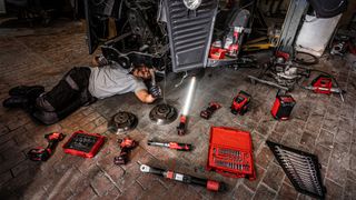 A man lying on the floor of a garage working on an engine and surrounded by Milwaukee tools
