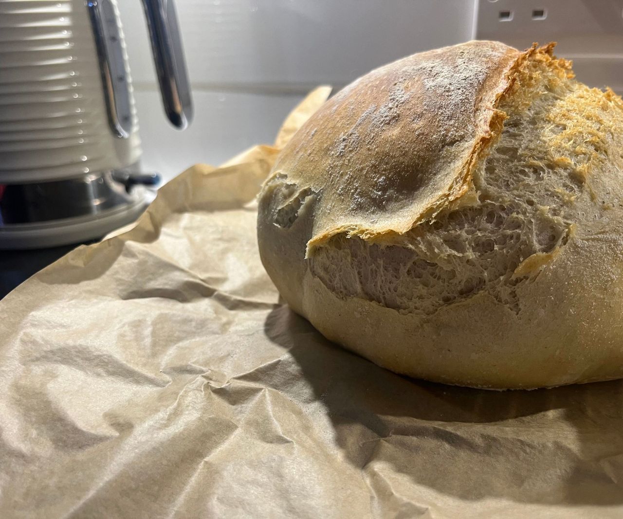 A fresh, browned loaf of bread on greaseproof paper on a cooling rack, viewed from the side, showing a bump in the shape of a pumpkin cocotte.