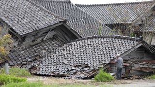 a man on his phone stands in front of a collapsed house, with the roof on the floor with grass in front