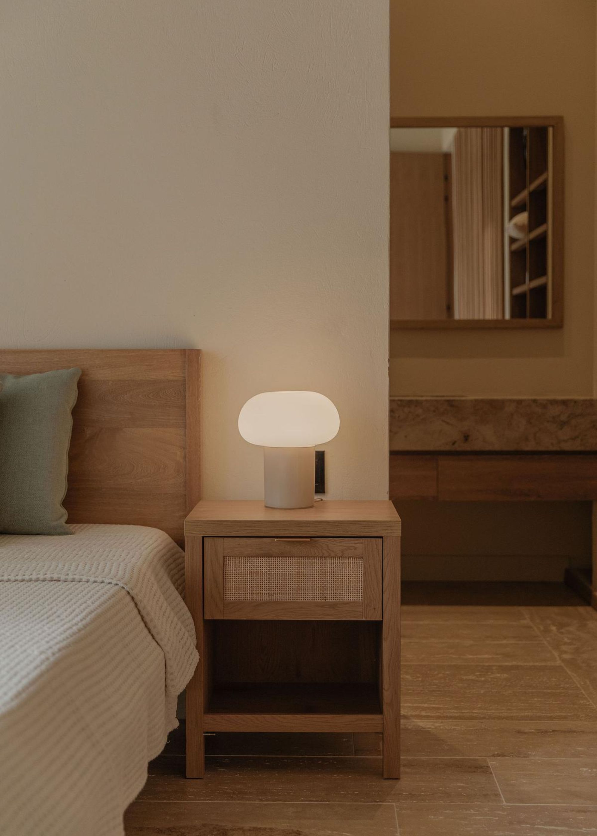 A beside table next to a double bed. On the table is a small table lamp that has been switched on.