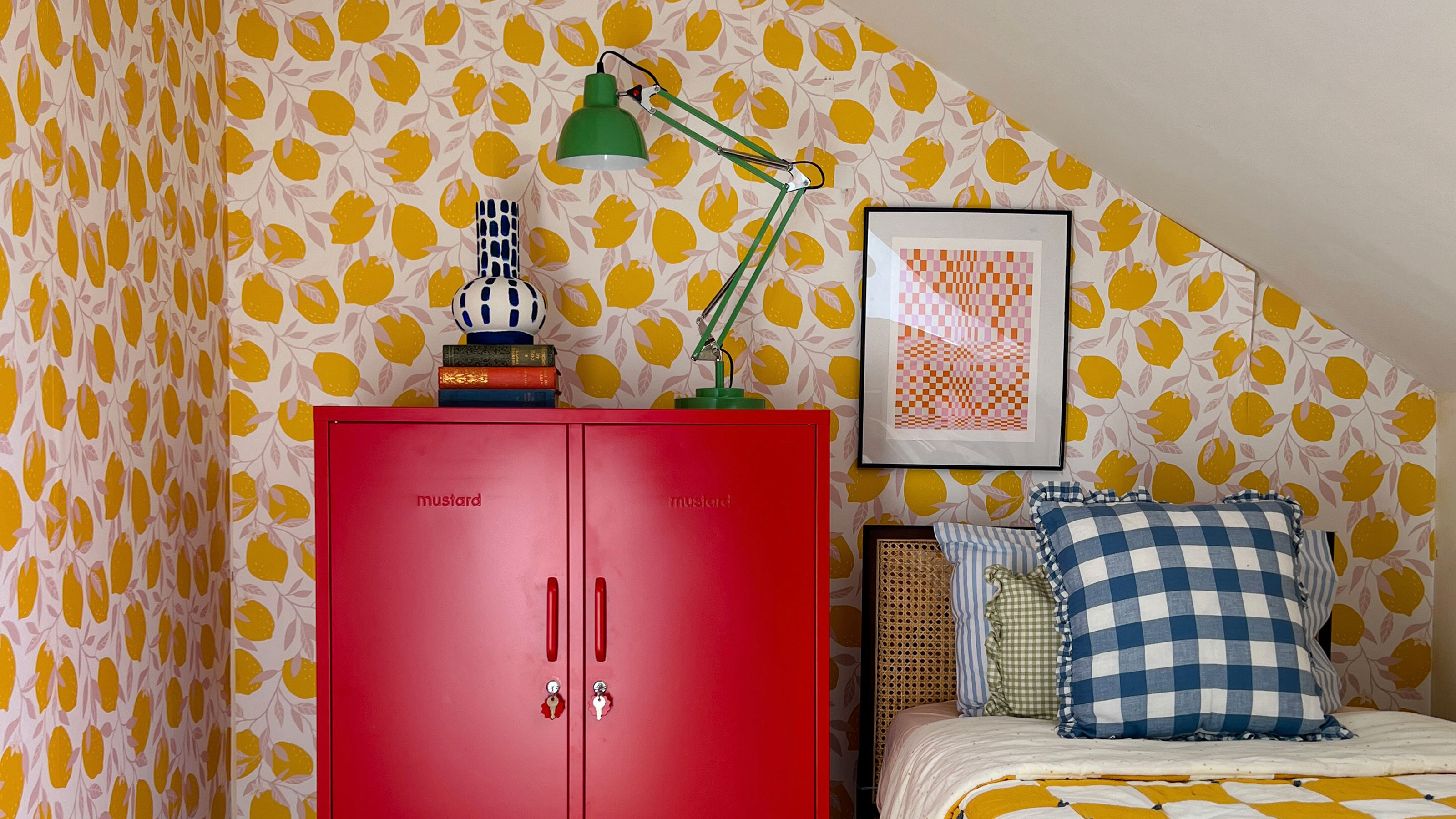 Eaves bedroom with yellow patterned wallpaper, and chequered yellow bedding. Red locker cabinet used as bedside table with green desk light