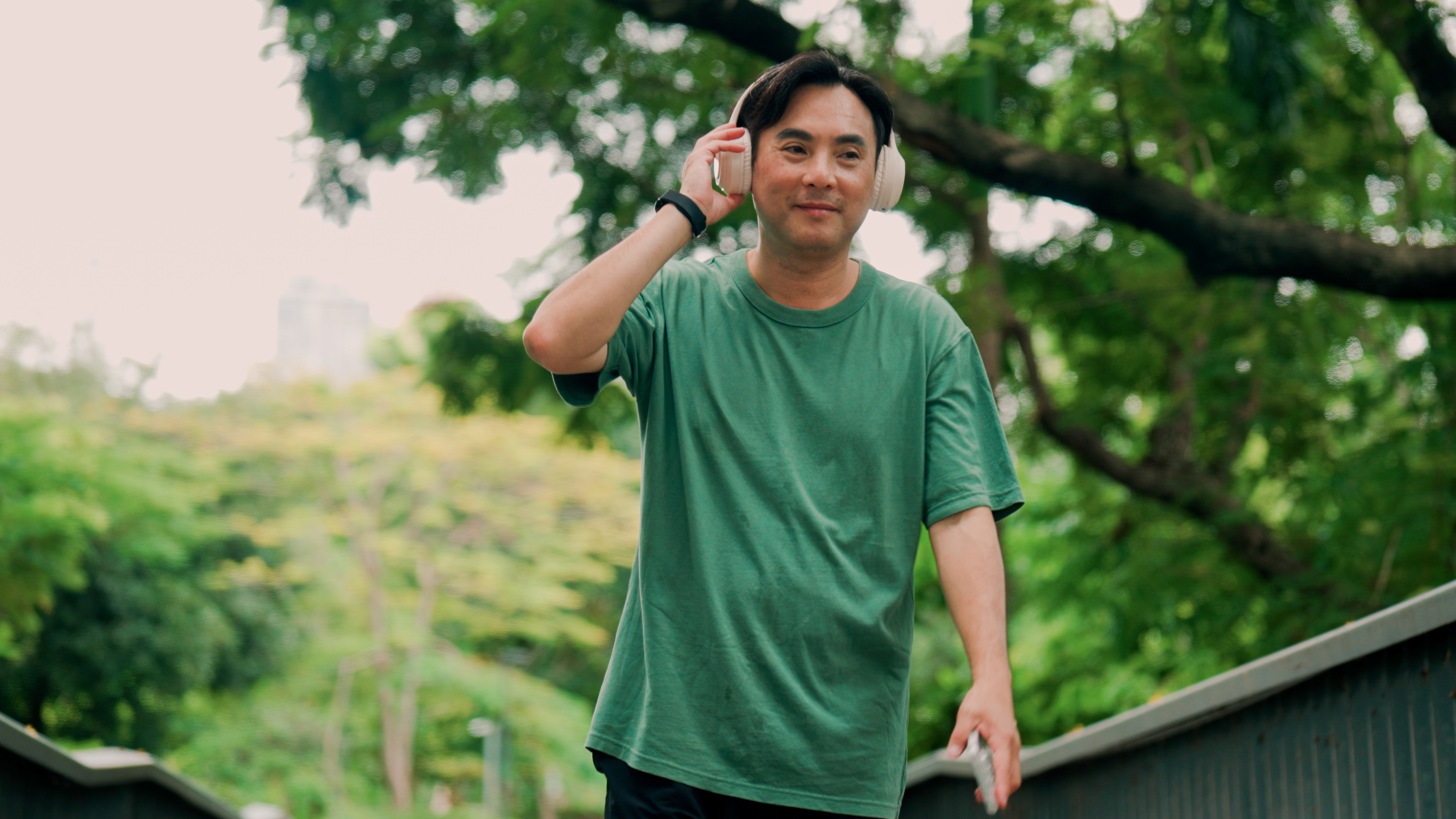A man in a loose-fitting t-shirt walks over a bridge adjusting his over-ear headphones.