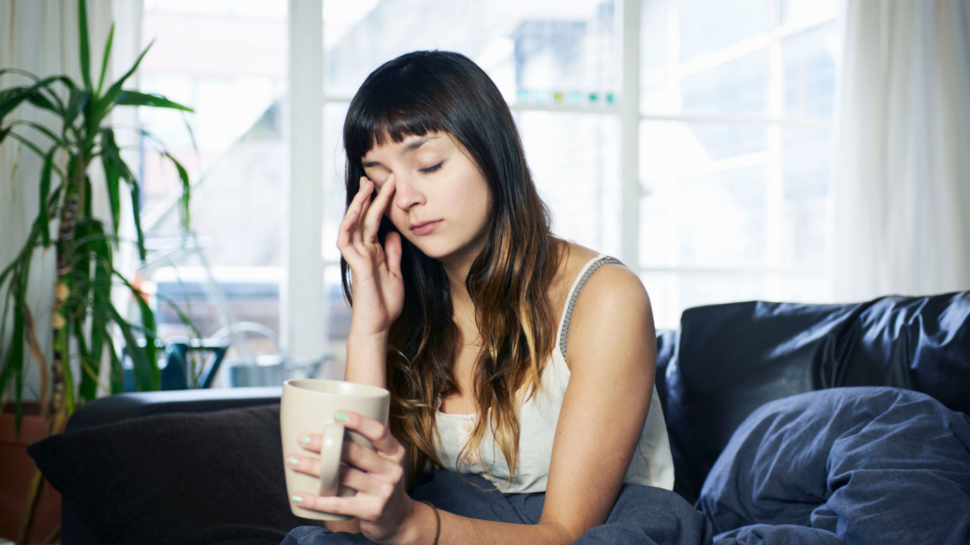 A woman sis on her couch looking tired, with her eyes closed. She has a cup of coffee in her hand.