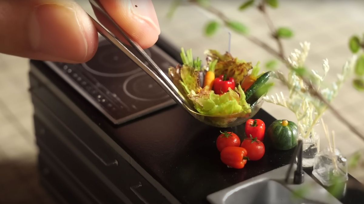 A shot of a miniature bowl of food on a diorama kitchen tip with a hand in frame using tweezers