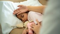 photo of a young girl napping with a stuffed rabbit as an adult checks her temperature with the back of their hand on her forehead