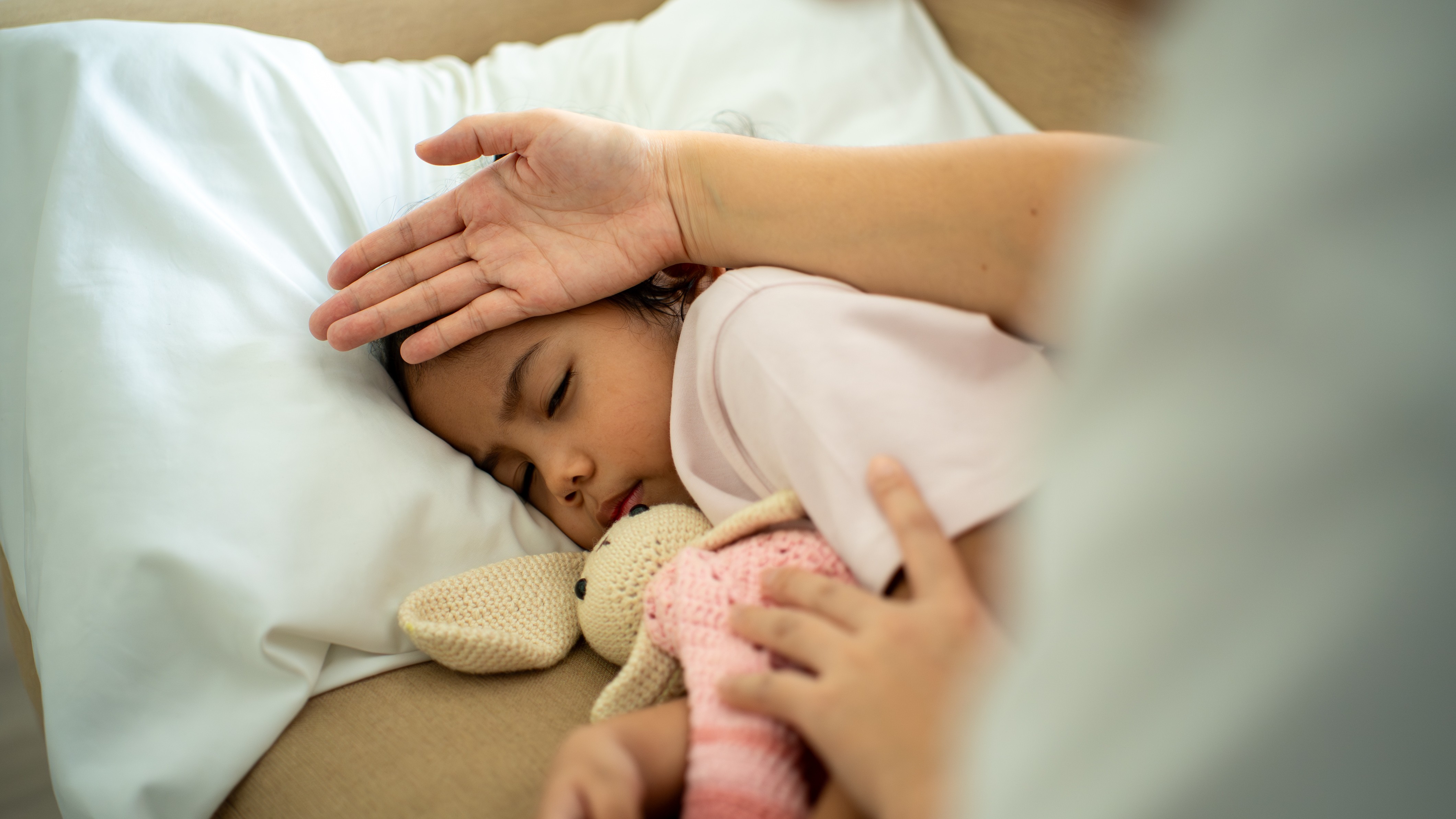 photo of a young girl napping with a stuffed rabbit as an adult checks her temperature with the back of their hand on her forehead