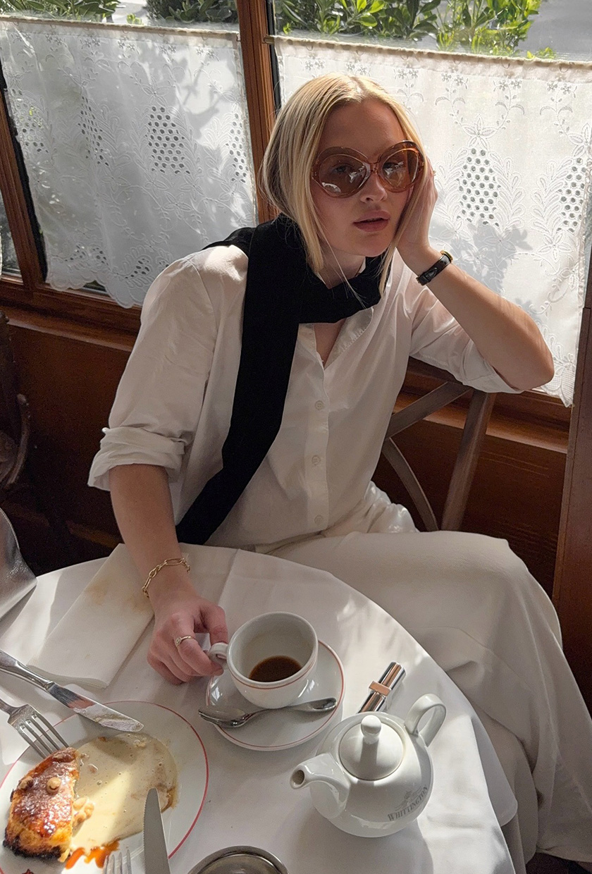 A woman in Paris wearing brown oversize '60s sunglasses with a black scarf, white button-down shirt, white trousers, and black sandals.