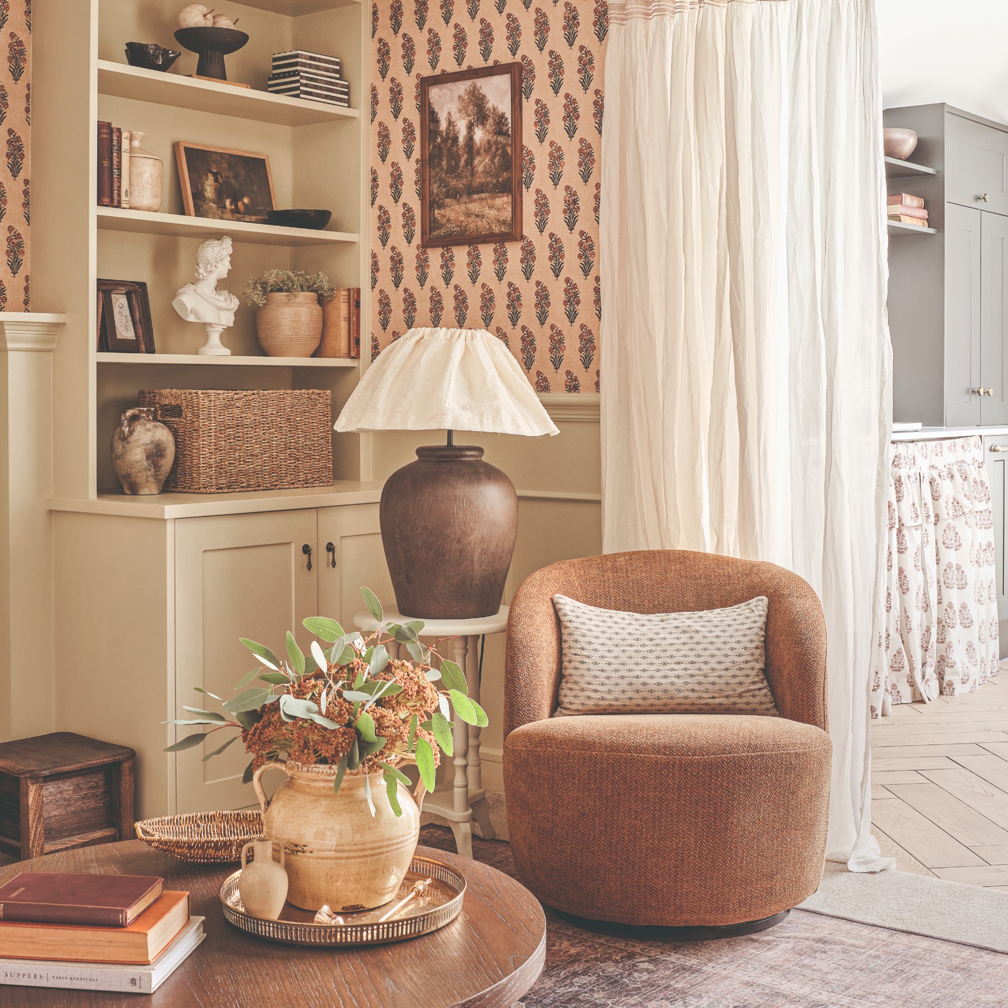 An open-plan living room with a curtain and an accent chair with a side table and large table lamp on top of it