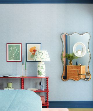 blue bedroom with blue walls and dark blue woodwork, colorful artwork, red bobbin console table and curvy mirror