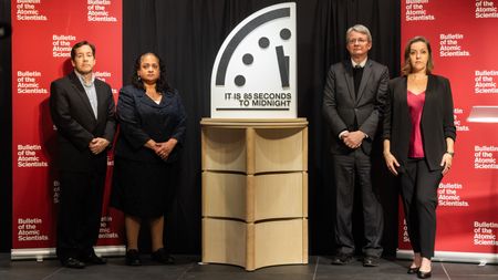 Four people stand solemnly on a stage on either side of the Doomsday Clock display during a press event for the Bulletin of the Atomic Scientists.