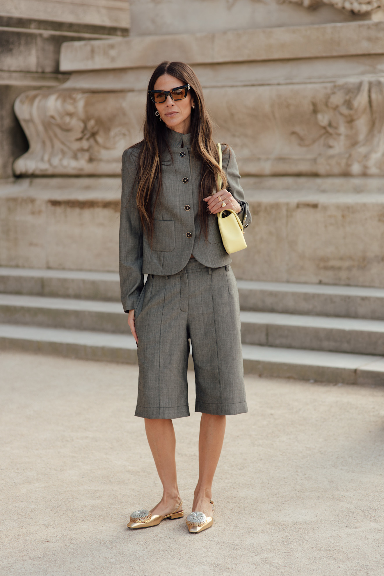Fashion week attendee wears grey blazer and bermuda shorts, gold flats, sunglasses, and yellow bag