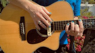 A photo of a man's hands on an acoustic guitar as he plays it. His fingers have acrylic nails with blue nail polish. 