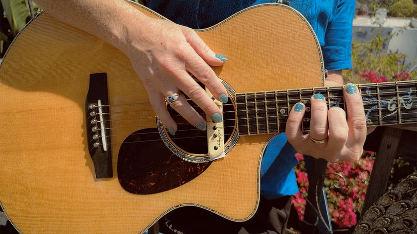 A photo of a man's hands on an acoustic guitar as he plays it. His fingers have acrylic nails with blue nail polish. 