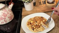 Plate of pancake next to stove with kettle on it. A hand holds a fork with a piece of the pancake on it