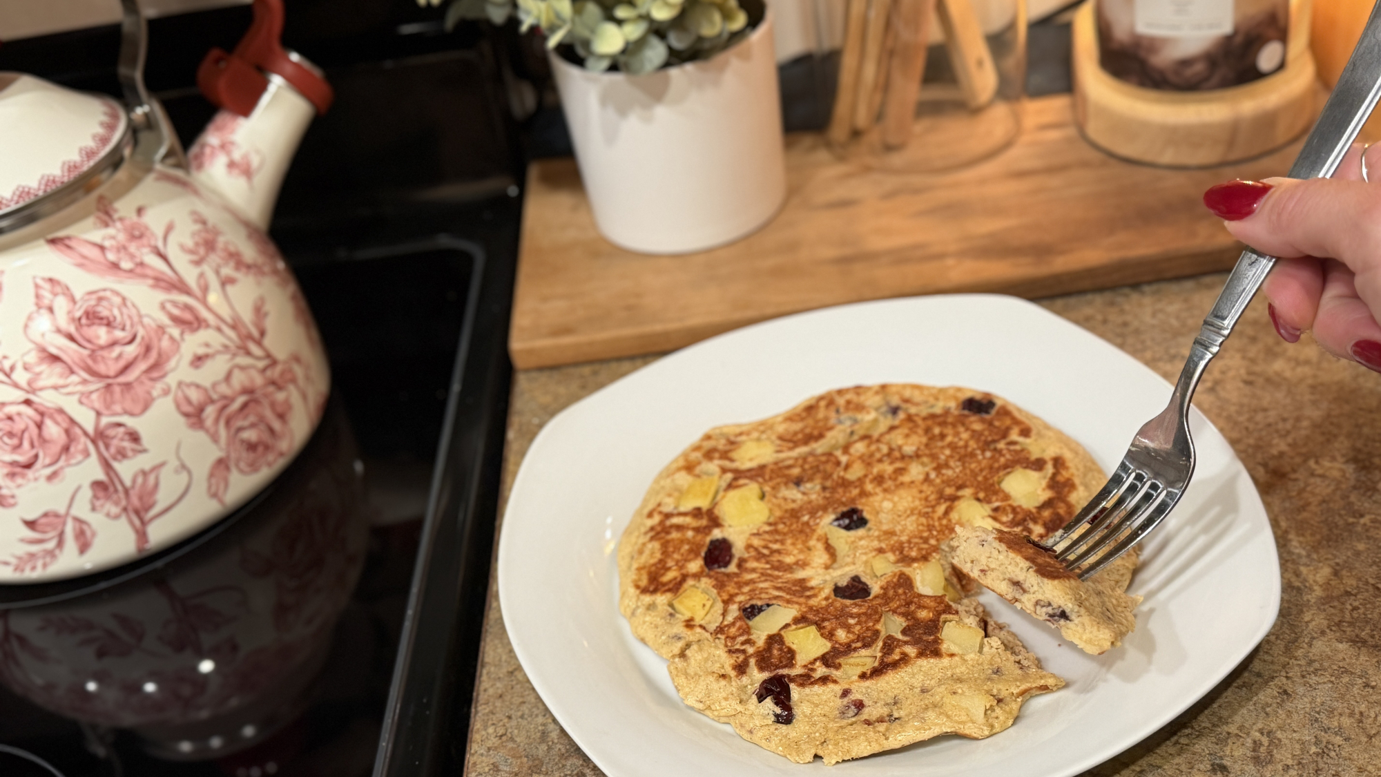 Plate of pancake next to stove with kettle on it. A hand holds a fork with a piece of the pancake on it