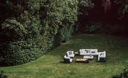Outdoor furniture including sofas and chairs upholstered in gray fabric by Potocco, photographed on grass in a garden surrounded by plants