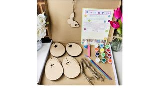 An activity box containing 2D cardboard eggs with a paint brush, a selection of different coloured paint pots and string to hang your eggs up with once they've been decorated