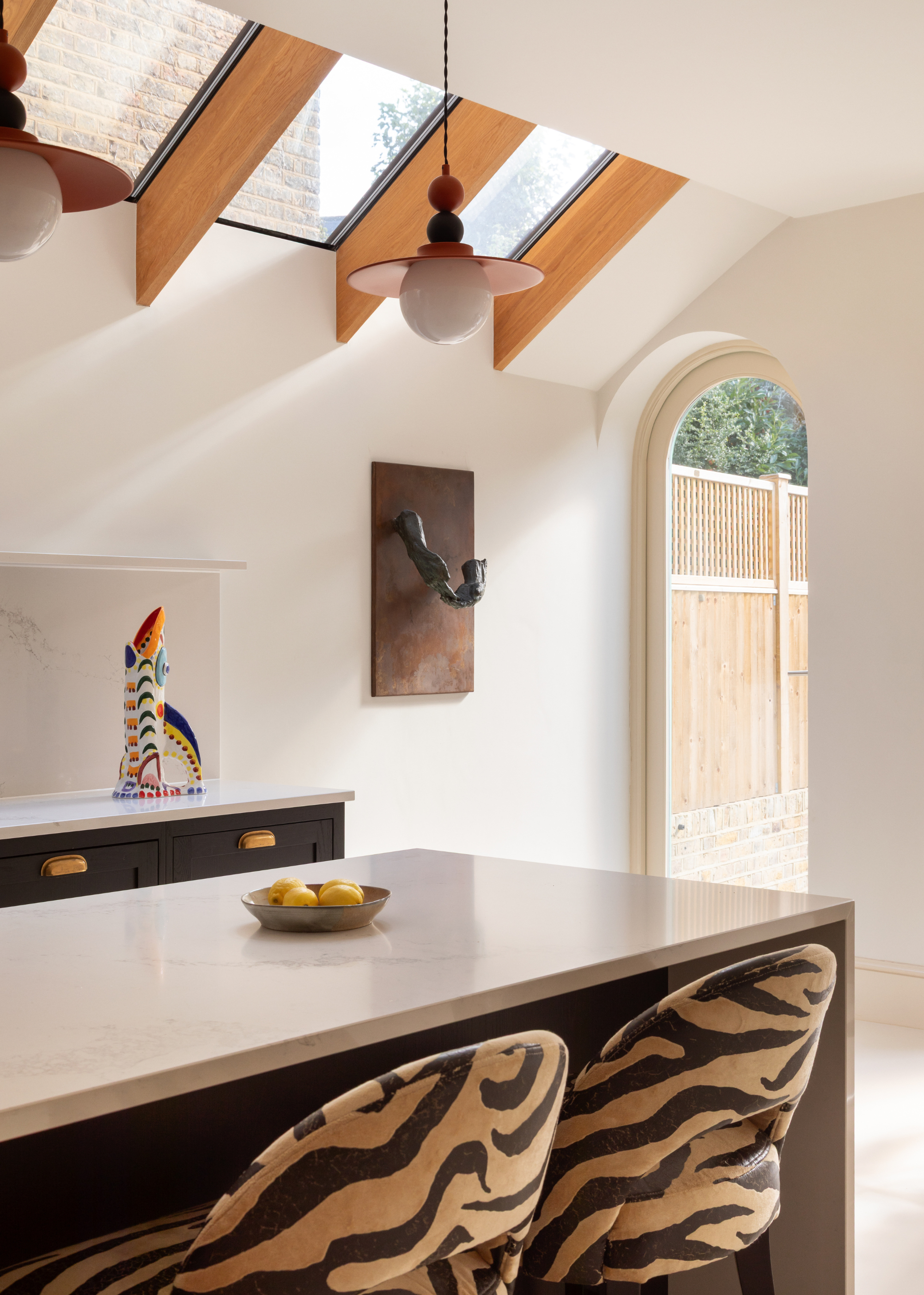 A bright white kitchen with slatted skylights, a stone isldn, a pair of tiger-print chairs, a bowl of lemons, and a colorful jug