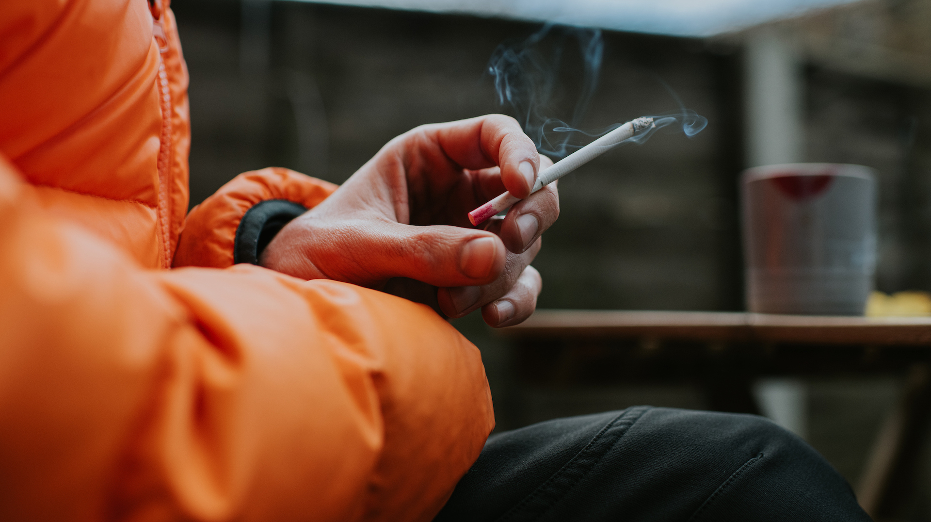A photograph of a person in a puffy jacket smoking a cigarette.