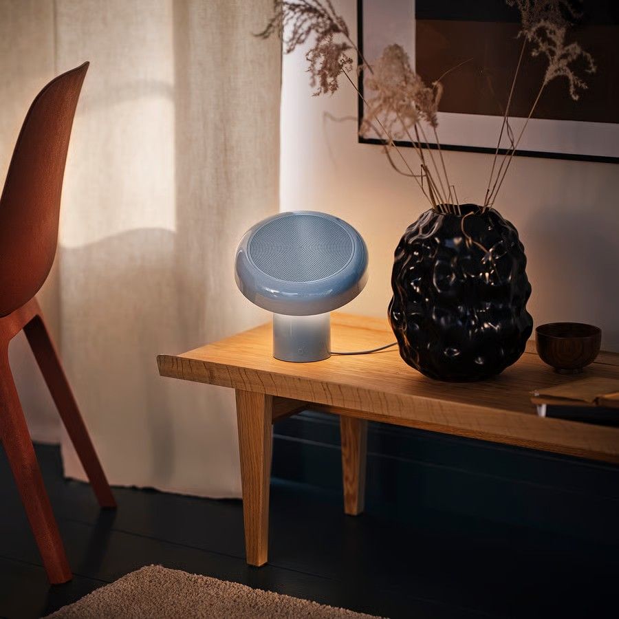 A baby blue Bluetooth speaker on a low, wooden bench next to a large black flower vase. The room is white with black floors, and there are white linen curtains drawn in the room.