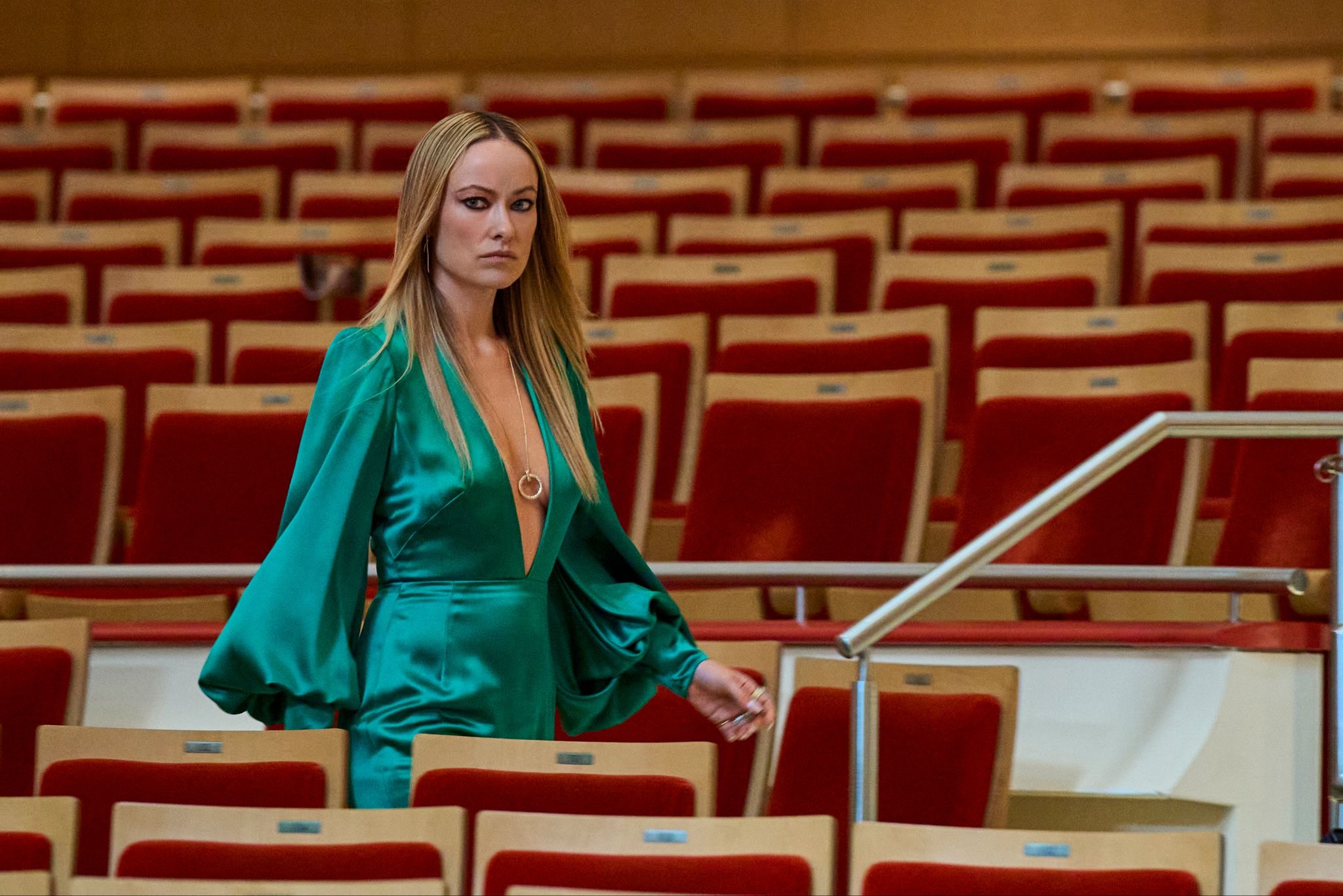 olivia wilde wears a teal low cut satin dress while walking through an empty auditorium in behemoth