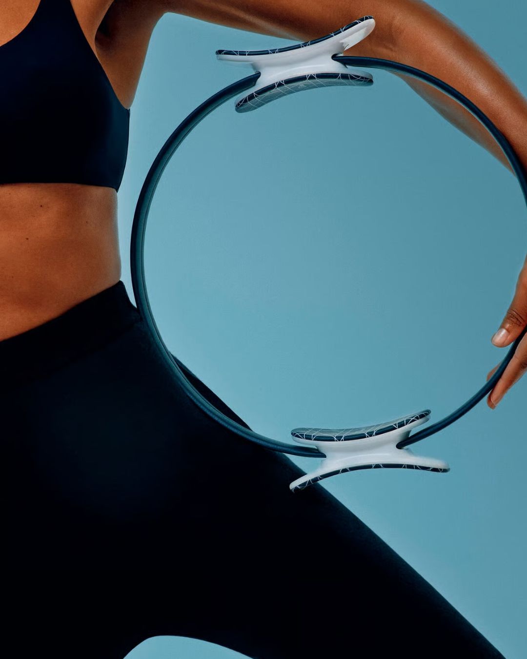 A Pilates ring, hold by a woman in a dark blue fitness set of leggings and cropped top, and shown against a pale blue background.