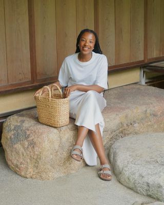 Female style influencer Taffy Msipa sits smiling on a large stone rock bench wearing a white t-shirt, white slip skirt, basket tote bag, and gray Birkenstock slide sandals.