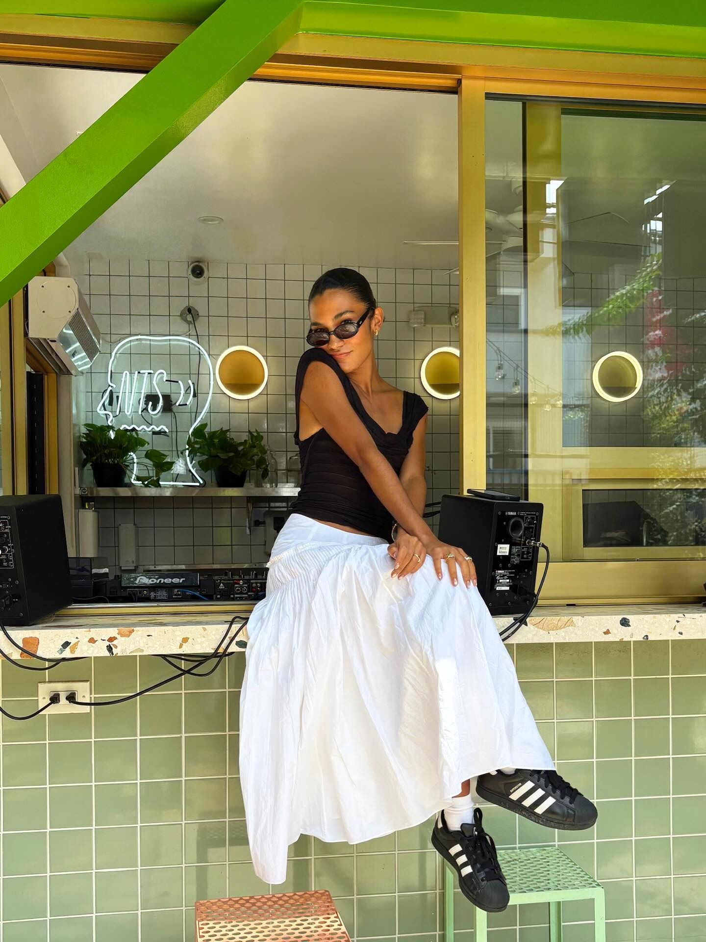Singer Olivia Dean sits on a tabletop wearing a white cotton skirt with Adidas trainers in black and a black sleeveless top.