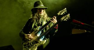 Ron 'Bumblefoot' Thal plays a doubleneck electric onstage in 2020. He wears a wide-brimmed hat and the stagelights are green-gray.