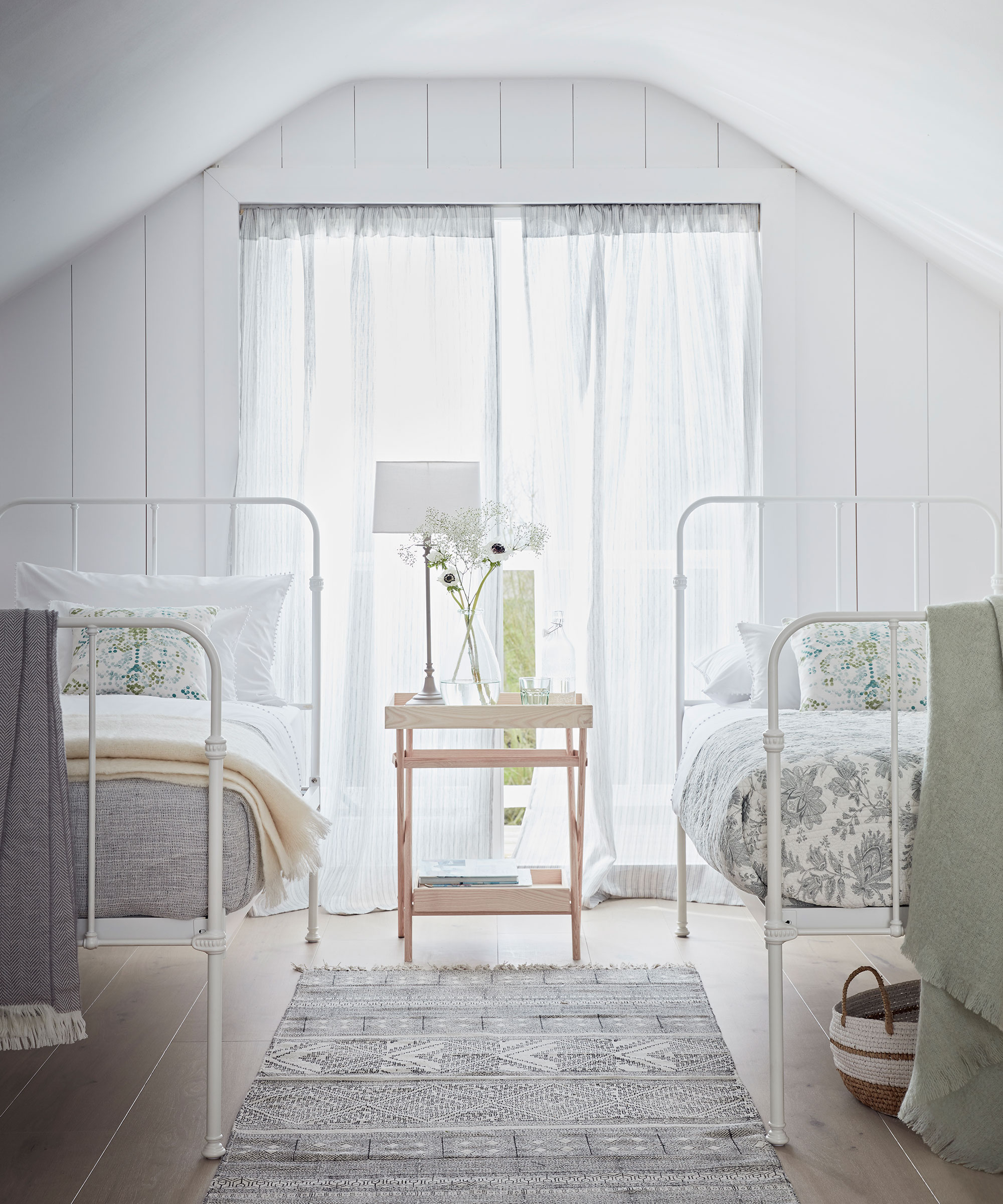 A light and airy white bedroom with two twin metal beds with toile curtains and white walls