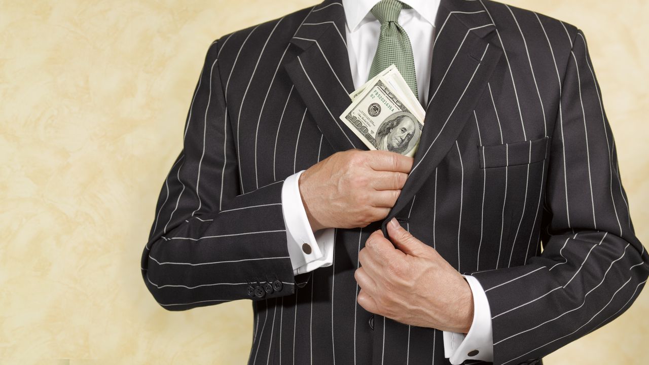 A man in a business suit slips a stack of hundred-dollar bills into an inside pocket of his suit jacket.