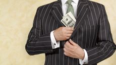 A man in a business suit slips a stack of hundred-dollar bills into an inside pocket of his suit jacket.