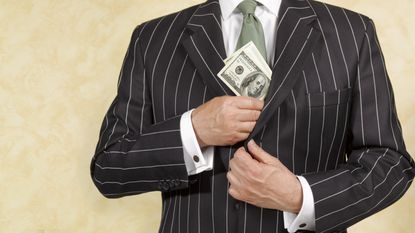 A man in a business suit slips a stack of hundred-dollar bills into an inside pocket of his suit jacket.