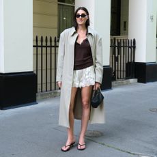 British style influencer Sarah Louise Blythe poses on a sidewalk wearing white satin lace-trimmed shorts, a trench coat, brown top, black oval sunglasses, mini black bag, and black flip-flop sandals