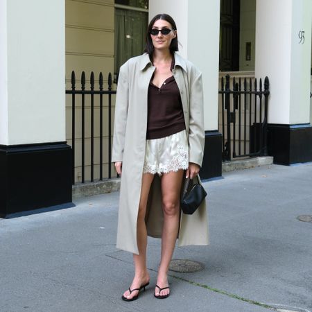 British style influencer Sarah Louise Blythe poses on a sidewalk wearing white satin lace-trimmed shorts, a trench coat, brown top, black oval sunglasses, mini black bag, and black flip-flop sandals