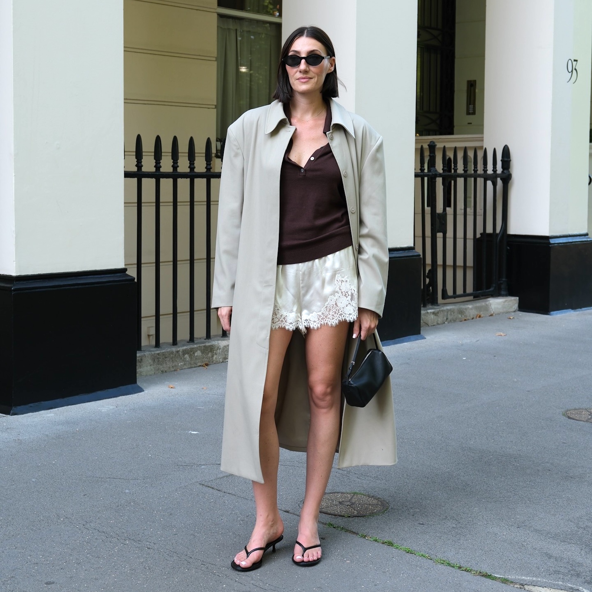 British style influencer Sarah Louise Blythe poses on a sidewalk wearing white satin lace-trimmed shorts, a trench coat, brown top, black oval sunglasses, mini black bag, and black flip-flop sandals