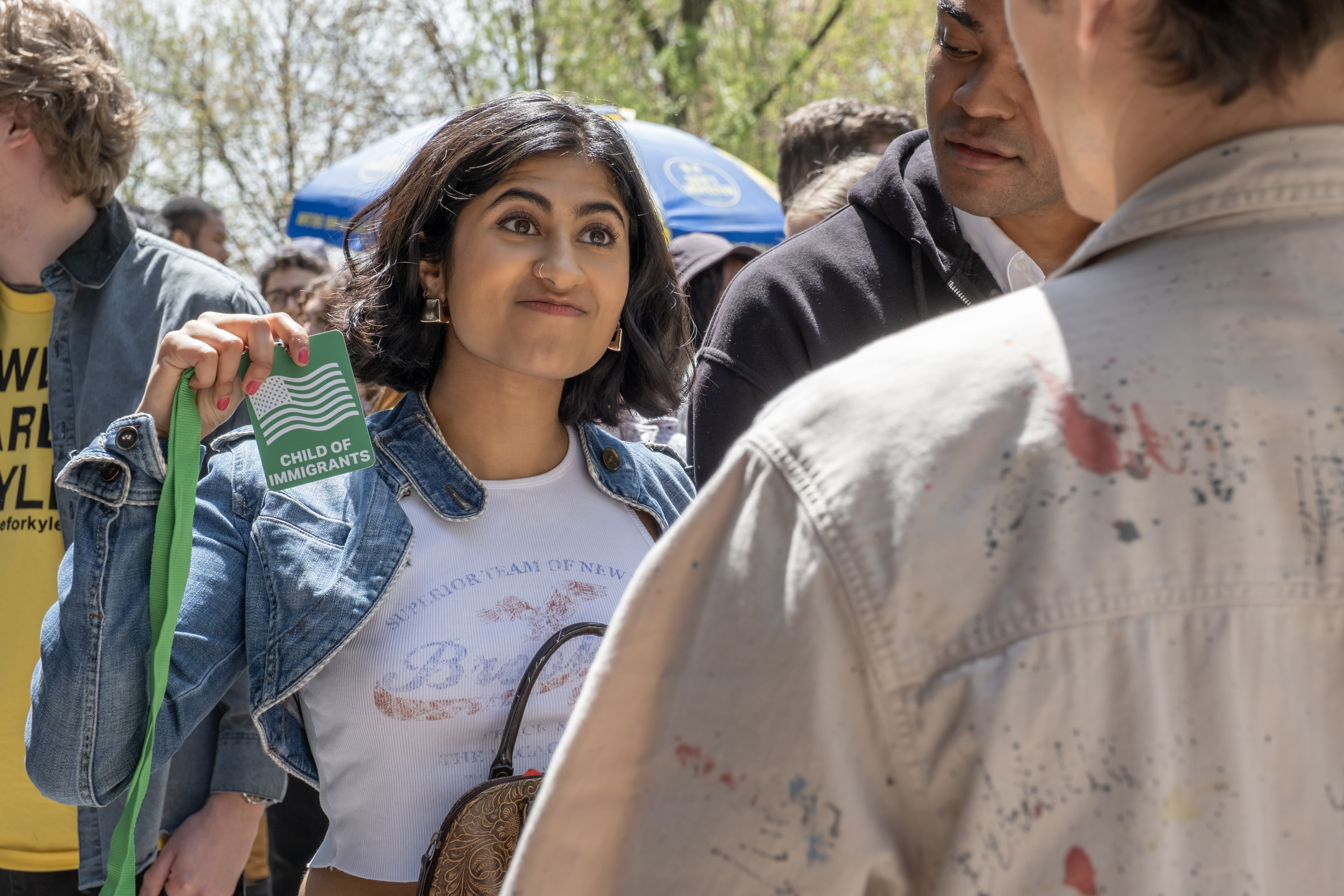 A woman (Amita Rao as Issa) holds up a green name tag reading "child of immigrants" as she stands in a crowd, in 'Adults.'