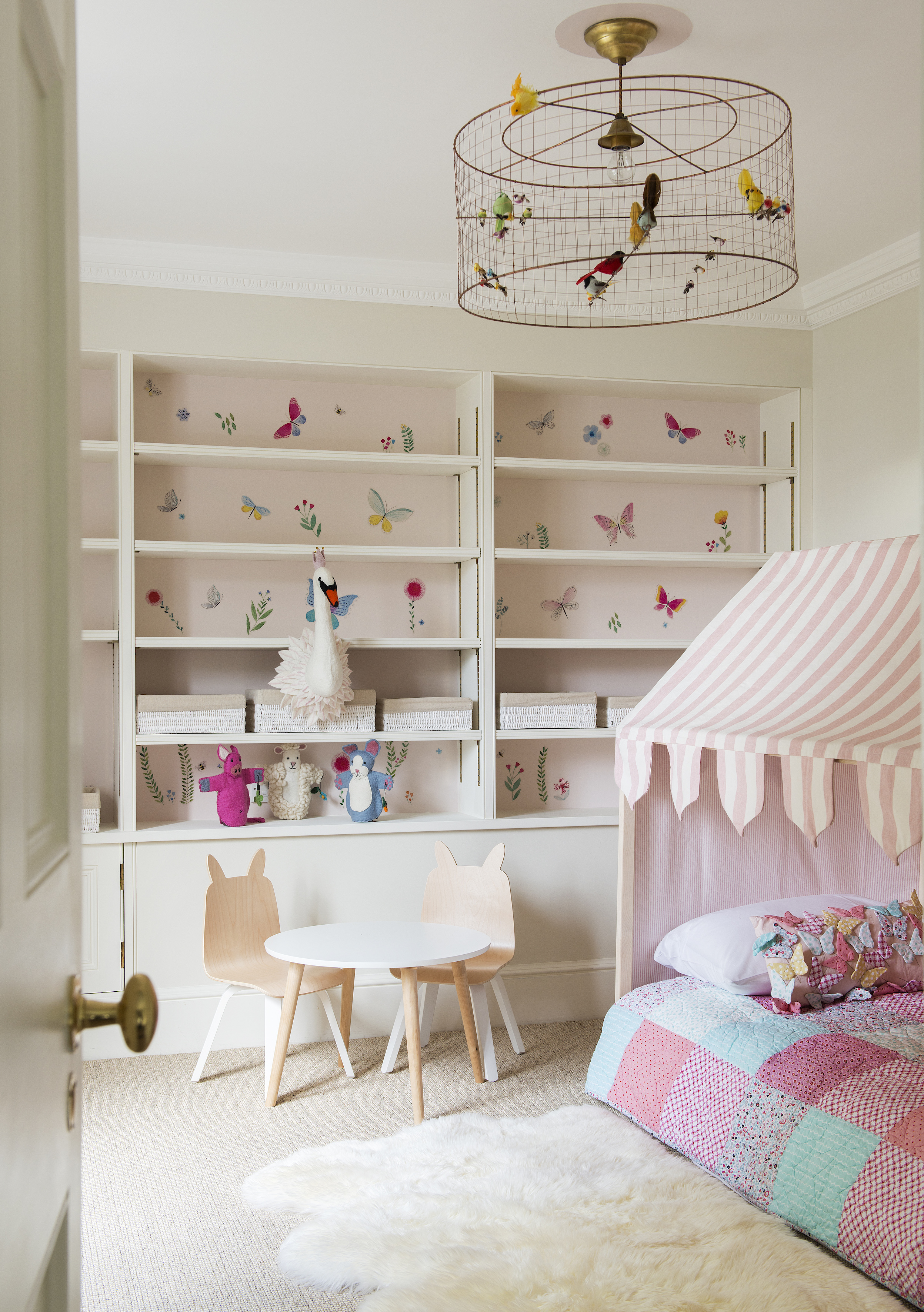 pastel pink girls bedroom with butterfly wallpaper behind shelves, bed with stripe tent style canopy, wire lampshade, table and chairs