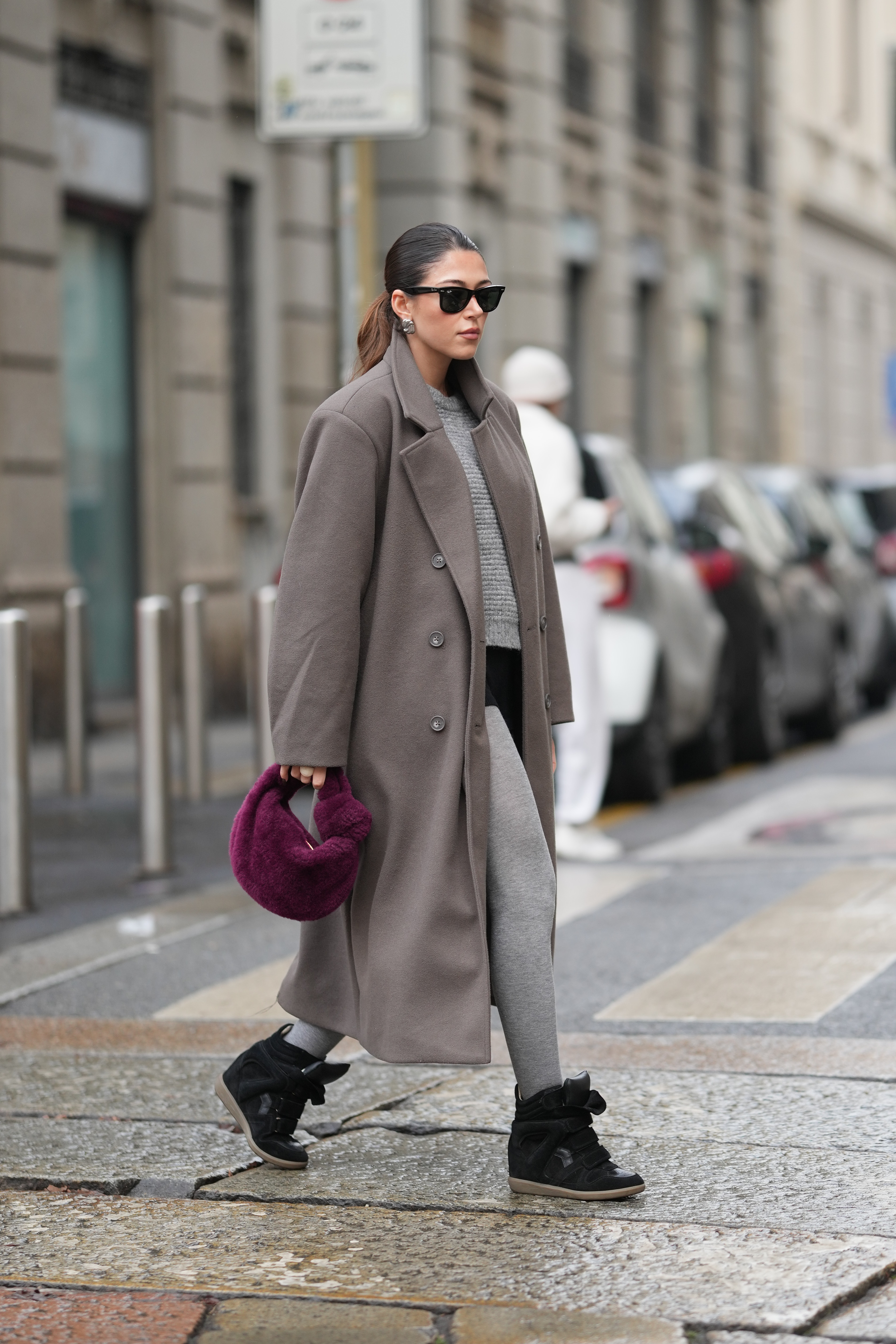 A guest wears heeled trainers and slicked-back dark hair pulled into a low bun, black sunglasses, silver hoop earrings, a deep plum faux-fur top-handle bag from Bottega Veneta, a taupe long wool coat,
