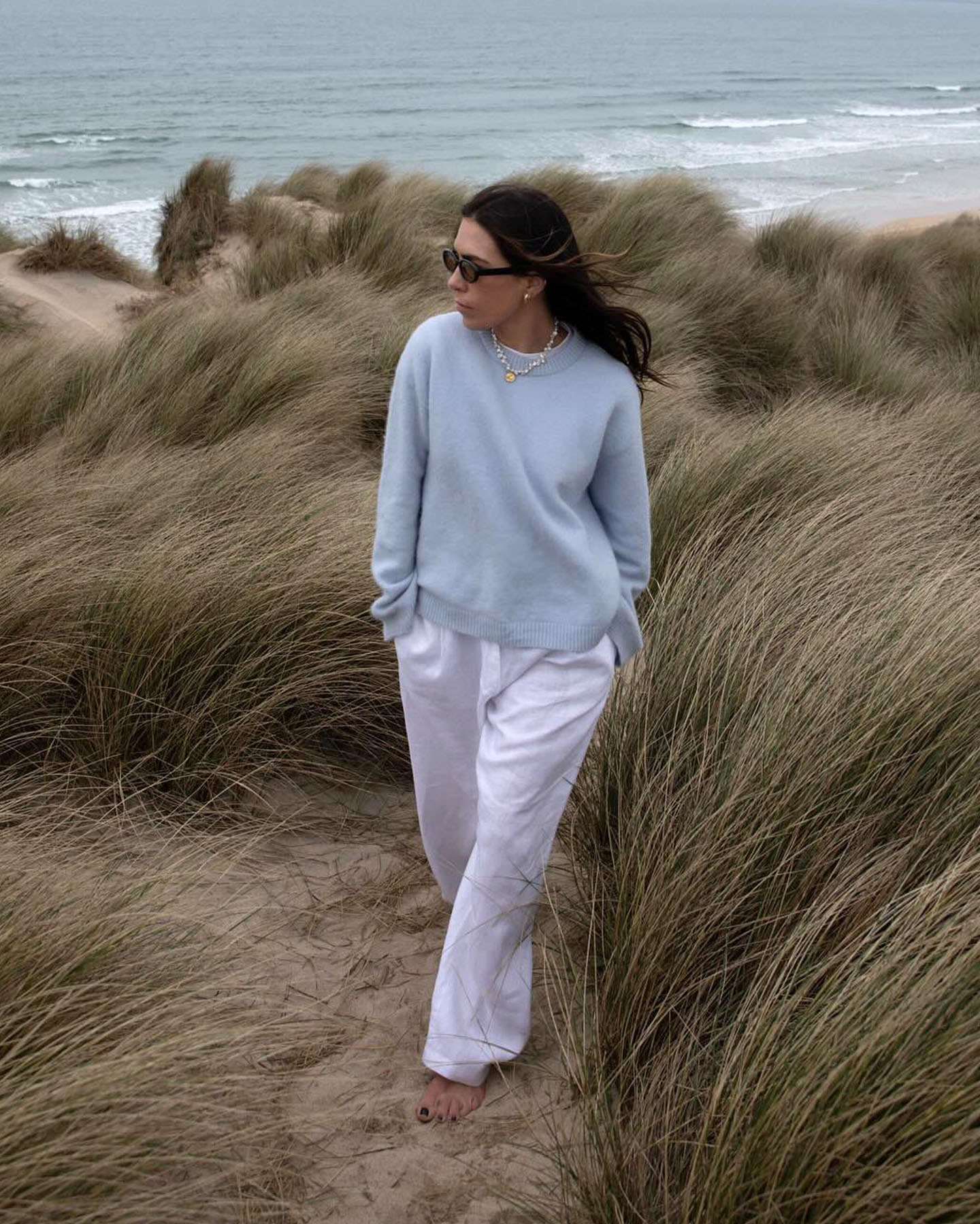fashion influencer Jessica Skye strolls on the beach wearing oval sunglasses, a light blue sweater, pearl necklace, and white linen pants