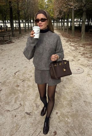 Black turtleneck outfit is shown in a photo of a French woman in the park wearing a brown stretchy headband with brown sunglasses and a black turtleneck layered under a gray crewneck mohair sweater styled with a matching mini shirt, black sheer tights, brown loafers, and a brown Hermes bag