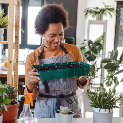 A woman in an apron smiles at a seed tray, surrounded by potted plants