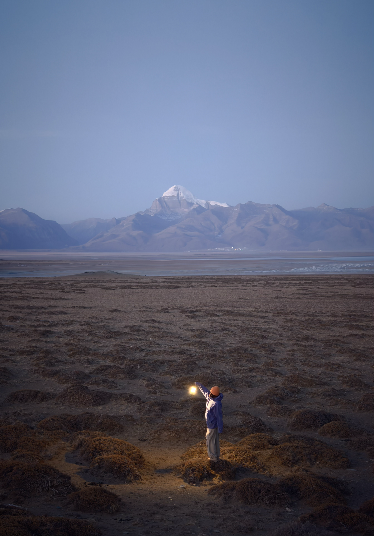 Person holding a lamp in the middle of a dimly lit field with a mountain in the background