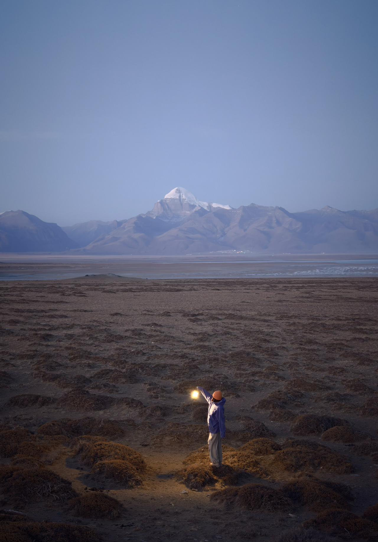 Person holding a lamp in the middle of a dimly lit field with a mountain in the background