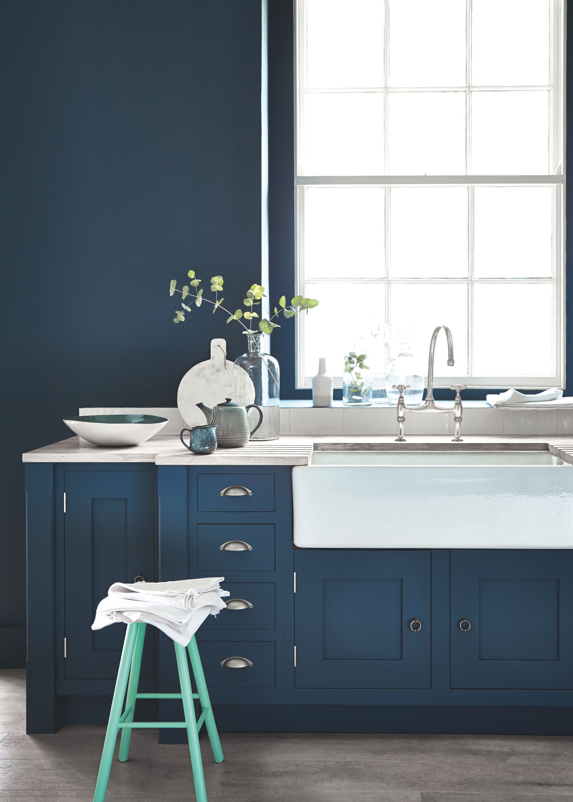 A kitchen with dark blue cabinets and walls, a Belfast sink and silver hardware, and a large traditional window.